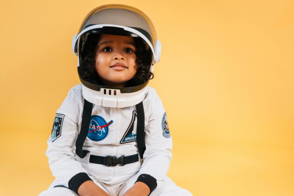 Adorable little African American girl in white space suit with helmet looking away while sitting against orange background