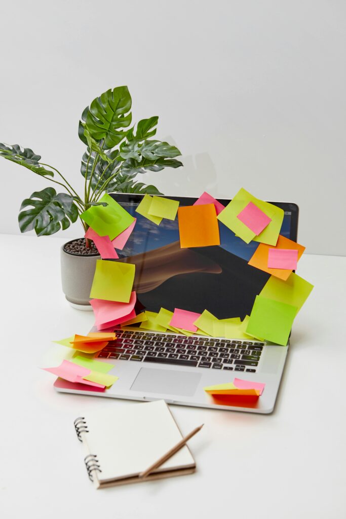 Laptop covered with colorful sticky notes, notebook, plant, and pencil on a minimalist desk.